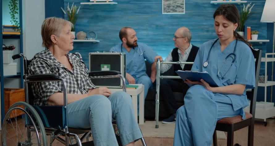 Retired woman with disability talking to nurse for medical checkup in nursing home. Healthcare assistant consulting elderly patient sitting in wheelchair for support and assistance.